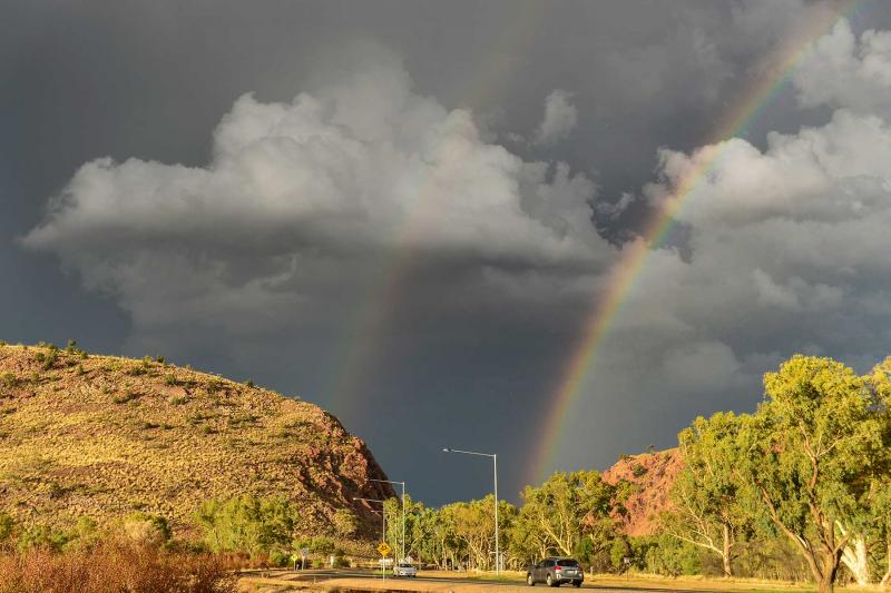 Sun shining on hills and trees with black clouds and rainbow in the background